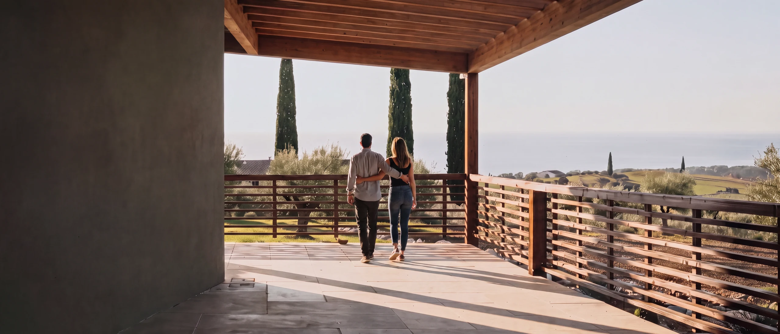 Two people walking away along a covered terrace. A slatted timber railing, cypresses and olive groves beyond it, and the sea in the far distance.